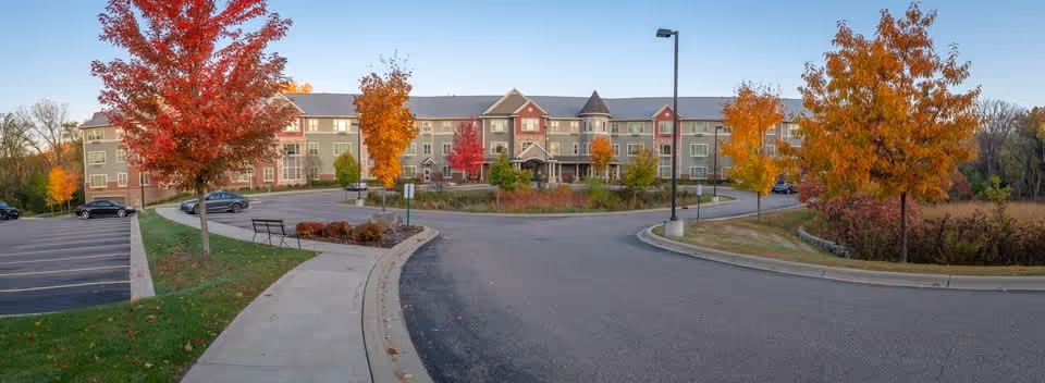 Front exterior view of The Shores of Lake Phalen senior living facility with a curved driveway, parking area, benches, and trees with autumn foliage.