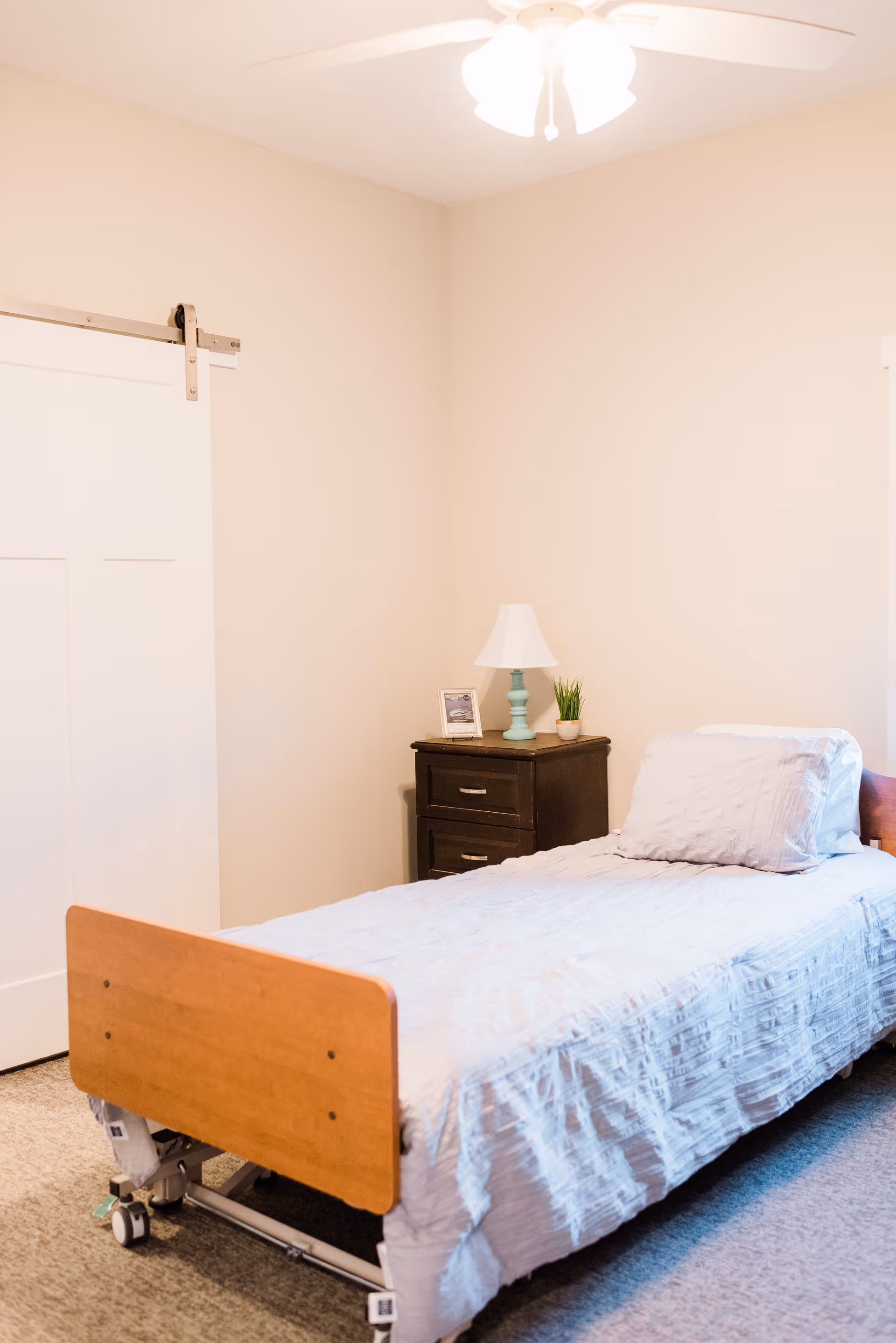 A simple bedroom with a single hospital-style bed with wooden headboard and footboard, white bedding, and a pillow. Next to the bed is a dark wooden nightstand with two drawers, a small lamp with a white shade, a framed photo, and a small potted plant. The room has beige walls, a white sliding barn door, and a ceiling fan with lights.
