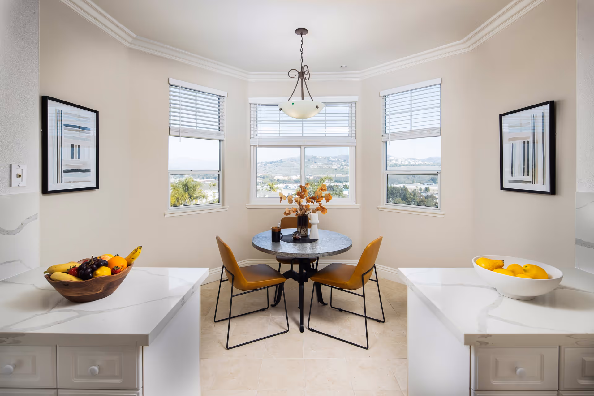 A bright dining nook with a round table and four mustard yellow chairs. The table has a decorative centerpiece with dried flowers and small items. The nook is surrounded by three windows with white blinds, offering a view of hills and trees outside. On either side of the nook are white marble countertops with bowls of fruit. Two abstract framed artworks hang on the walls.