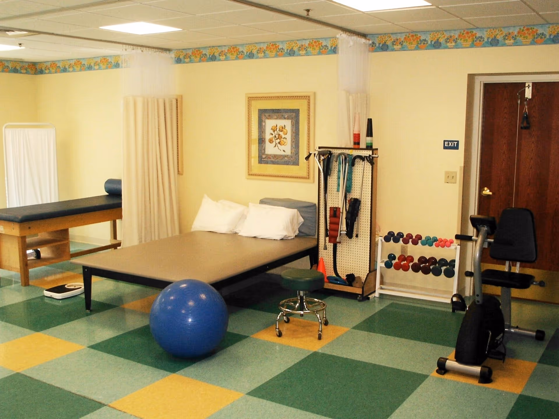 A rehabilitation room with a therapy bed, exercise ball, small stool, stationary exercise bike, and a rack holding various exercise bands and small dumbbells. The room has a tiled floor with green, yellow, and blue squares, light yellow walls with a floral border near the ceiling, and a framed picture on the wall.