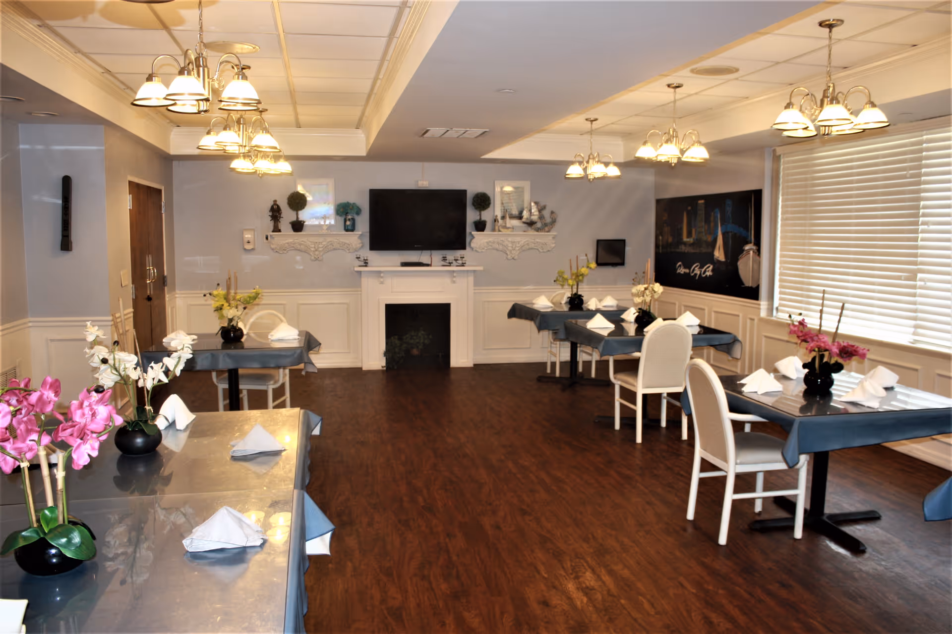 A dining room with several tables covered with blue tablecloths, each set with white napkins and black vases holding pink and white flowers. The room has wooden flooring, white wainscoting on the walls, and multiple hanging light fixtures. A flat-screen TV is mounted above a white fireplace on the far wall, with decorative shelves on either side. Large windows with blinds are on the right side of the room.