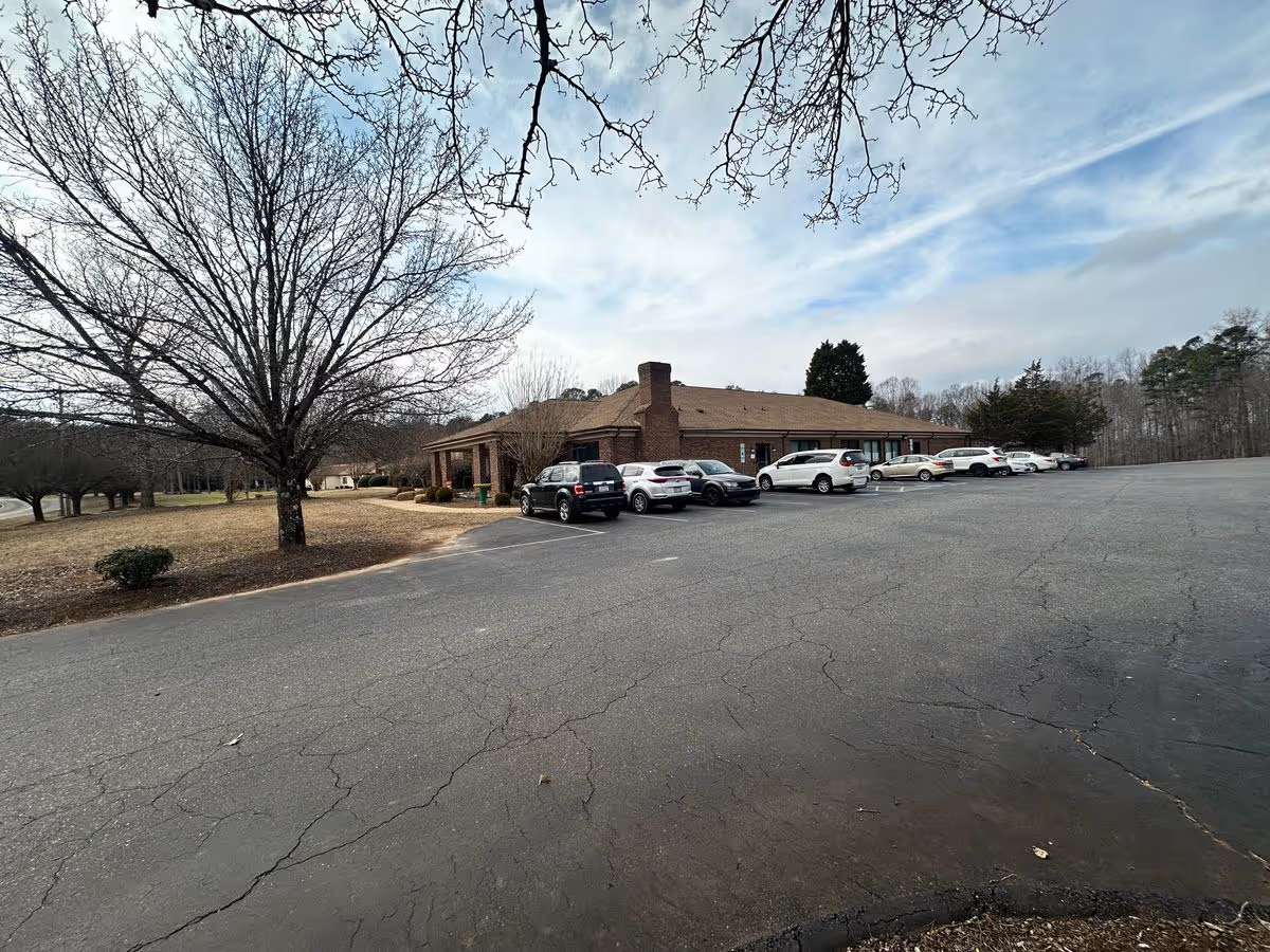 One-story brick senior living building with parked cars in a large cracked asphalt lot and leafless trees under a cloudy sky.