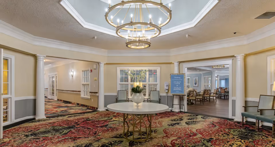 Spacious circular lobby with a central round table and vase beneath a tiered chandelier, flanked by columns and seating with patterned carpet leading to adjoining rooms.