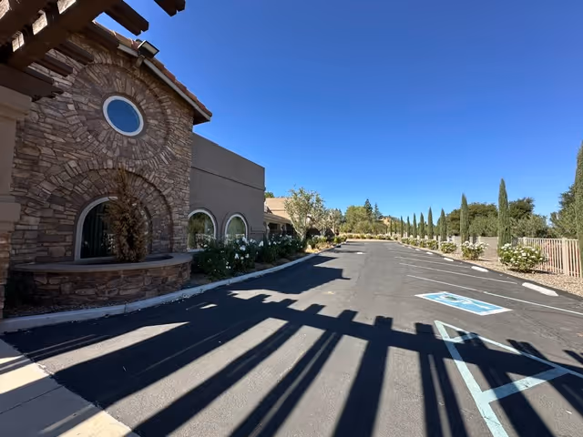Exterior view of a building with stone and stucco facade under a clear blue sky. The building has circular and arched windows, and there is a parking lot with marked handicapped parking spaces. Trees and shrubs line the parking area.