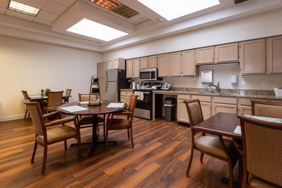 A communal kitchen and dining area with wooden floors, beige cabinets, stainless steel appliances including a refrigerator, microwave, and stove. There are several tables with chairs arranged around them, and the room is lit by overhead skylights.