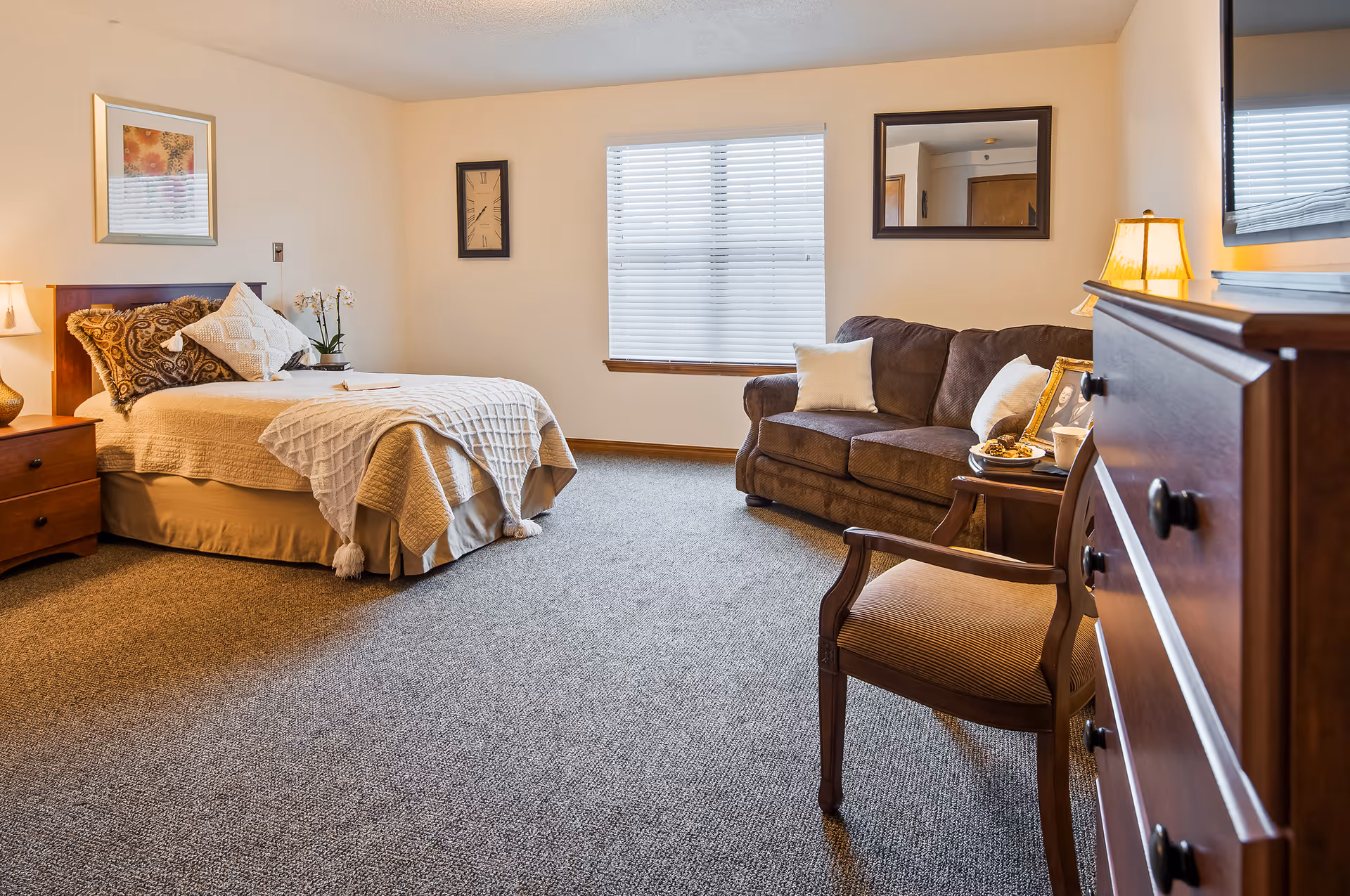 A cozy bedroom in Asbury Village featuring a neatly made bed with decorative pillows and a throw blanket, a wooden nightstand with a lamp, a brown sofa with white pillows, a wooden chair, a dresser with a lamp and framed photos, a window with blinds, a wall clock, and a framed mirror.