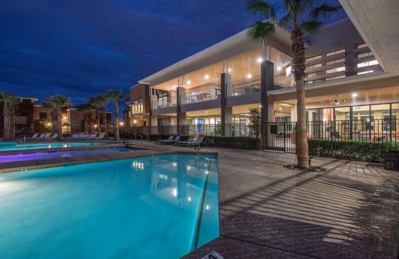 Illuminated outdoor swimming pool and lounge area at night in front of a modern two-story building with palm trees.