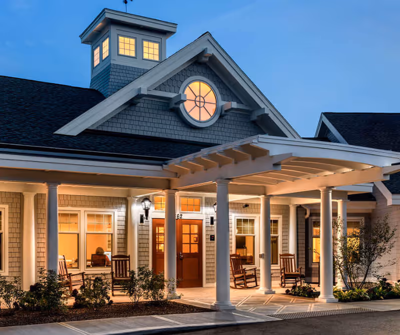 Front entrance of a well-lit senior living building at dusk with a covered porch, columns, rocking chairs, and illuminated windows.