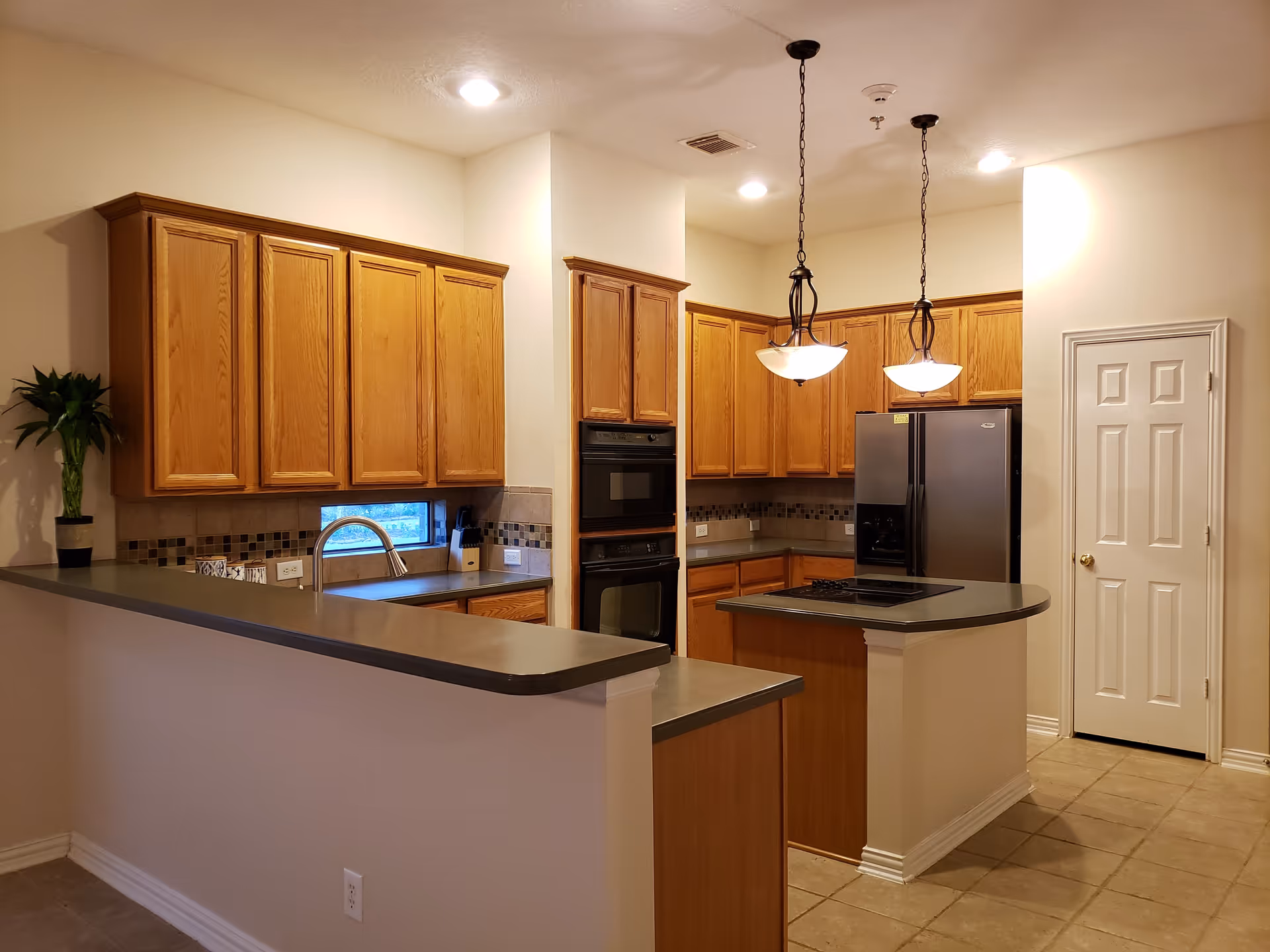 Open kitchen with oak cabinets, a central island with cooktop, stainless steel refrigerator, pendant lights, and a bar counter.