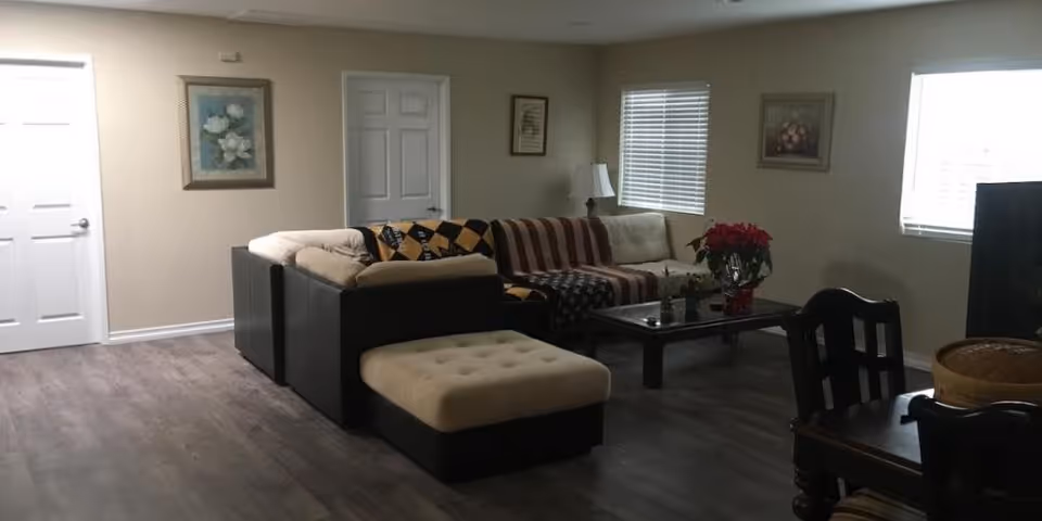A cozy living room with a sectional sofa covered with blankets, a coffee table with a red poinsettia plant, two framed paintings on beige walls, a window with blinds, a floor lamp, and a wooden dining table with chairs in the foreground.