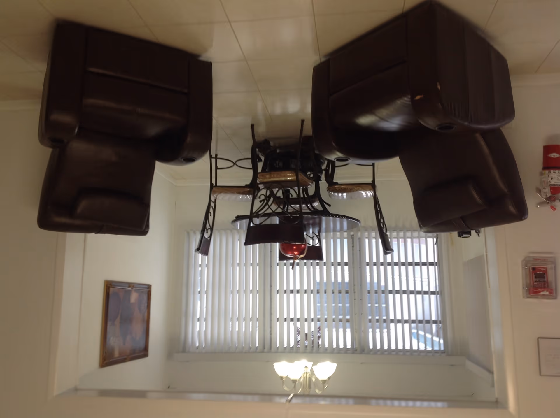 A small dining area with a round glass-top table and four metal chairs with cushioned seats. Two brown leather recliners are positioned on either side of the table. The room has white walls, vertical blinds covering a window, a framed picture on one wall, and a ceiling light fixture. A fire extinguisher and fire alarm are mounted on the wall near the window.
