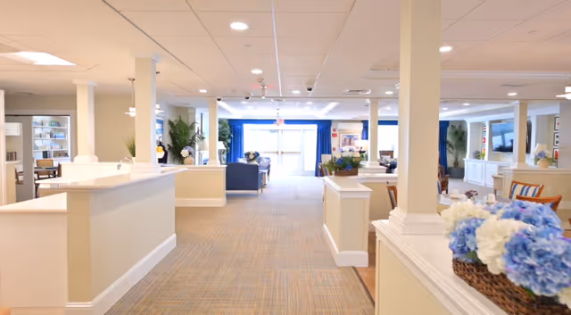 Bright and spacious senior living facility common area with beige walls and carpeted floor. The room features white columns, a reception desk on the left, seating areas with blue and wooden chairs, and decorative plants and flowers. Large windows with blue curtains allow natural light to fill the space.