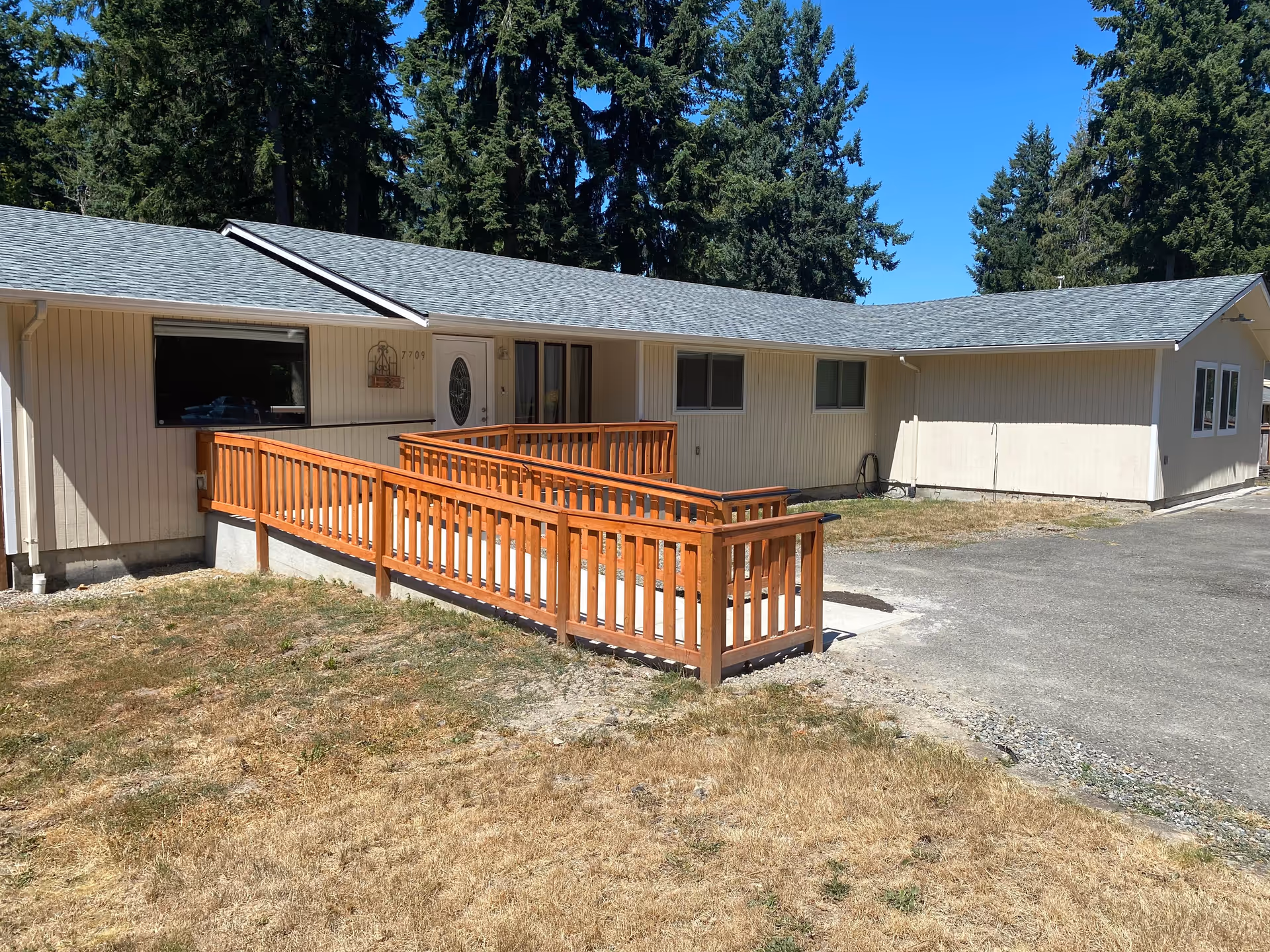 Exterior view of a single-story beige building with a gray shingled roof, featuring a wooden wheelchair-accessible ramp leading to the front door. The building is surrounded by dry grass and tall evergreen trees under a clear blue sky.
