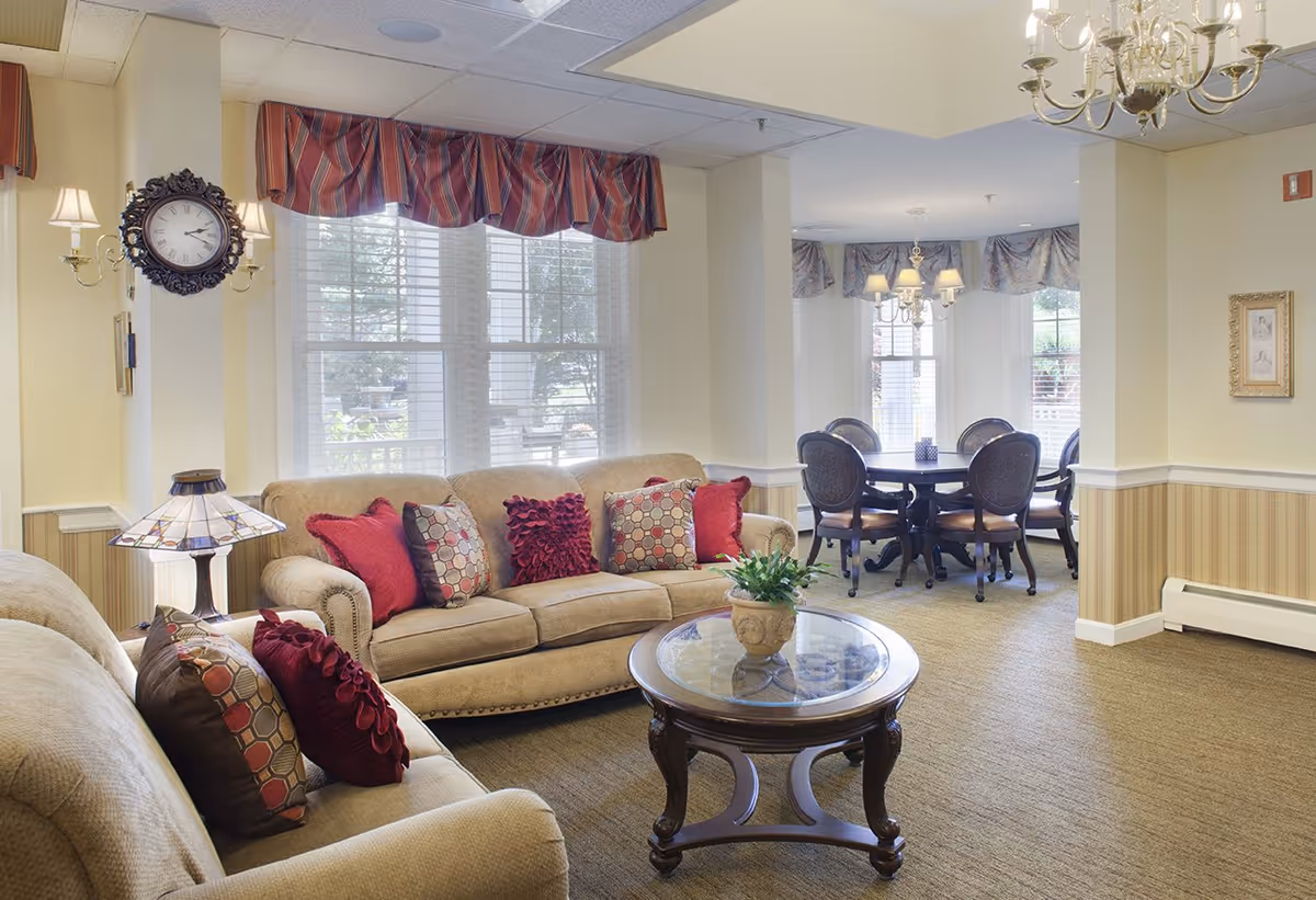 A bright living room with beige sofas and red decorative pillows around a glass-top coffee table, with a dining table and large windows in the background.