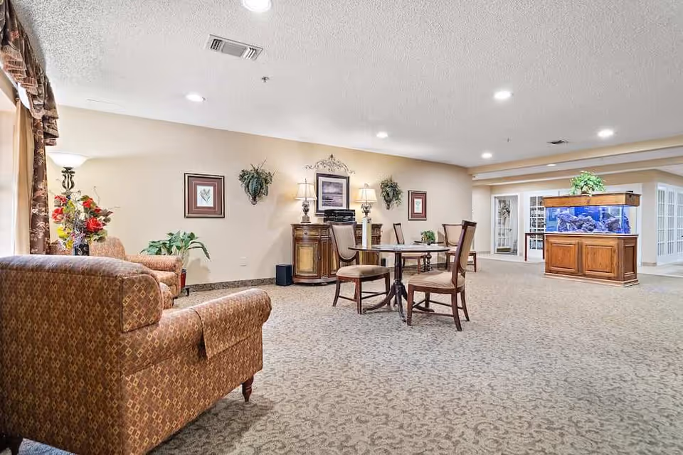 A spacious and well-lit common area in a senior living facility featuring patterned carpet, a round table with four chairs, two upholstered armchairs, decorative plants, framed artwork on the walls, a wooden cabinet with lamps, and a large aquarium with blue lighting on a wooden stand.