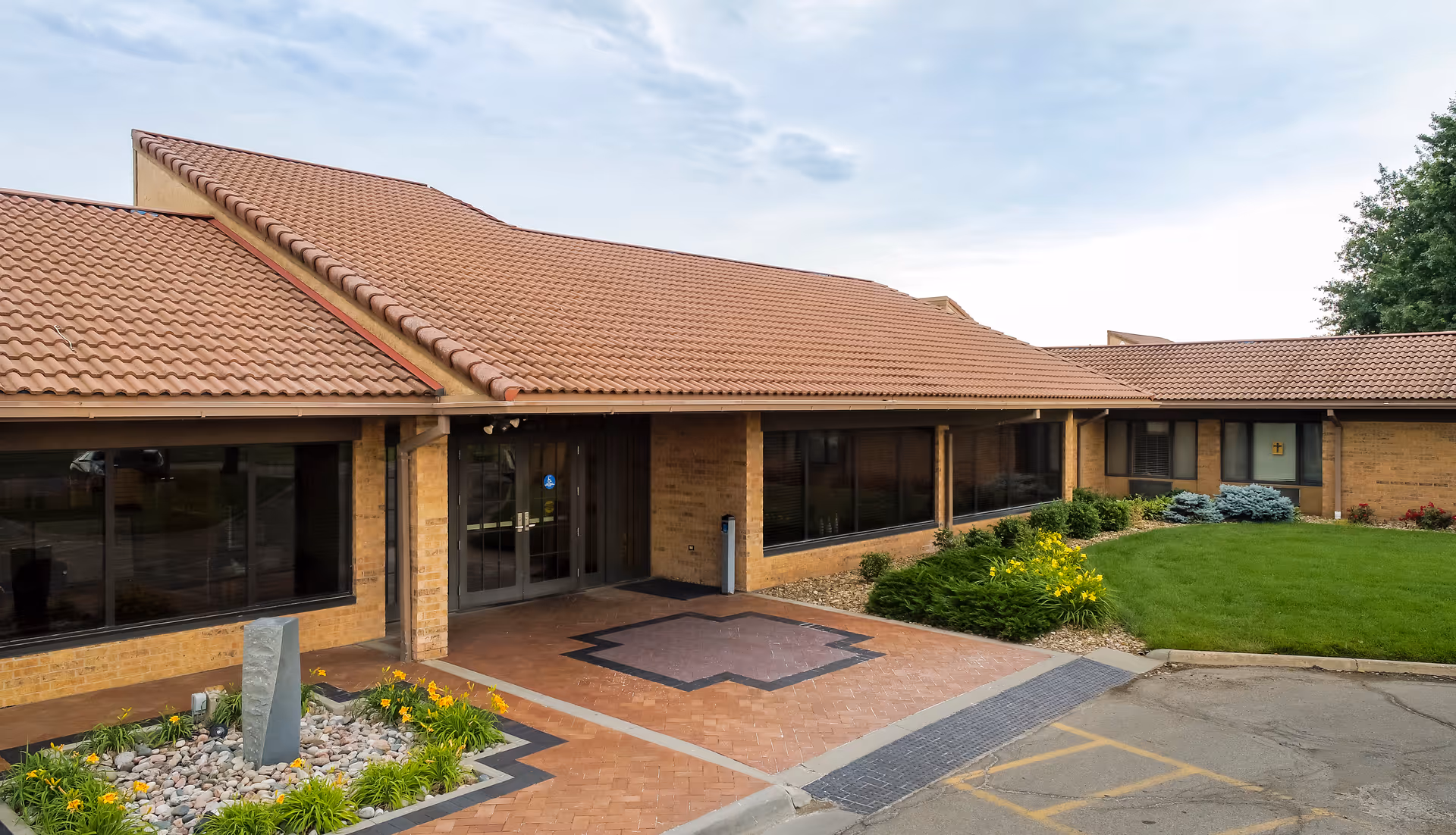Exterior view of Rolling Hills Health & Rehab of Topeka showing a single-story building with a tiled roof, large windows, a brick facade, landscaped garden with green grass, yellow flowers, and a stone feature near the entrance.