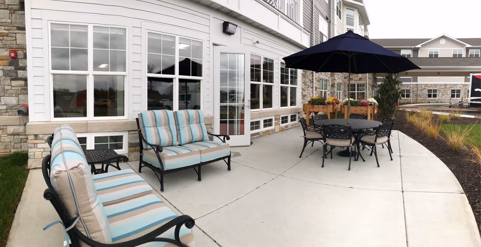Outdoor patio area at a senior living facility with cushioned metal chairs and a loveseat with striped cushions, a round metal table with an umbrella, and flower boxes near the building's windows and door.