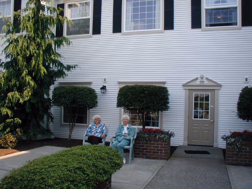 Two elderly women sitting on chairs outside a white building with large windows and neatly trimmed bushes, with flower planters on either side of a beige door.