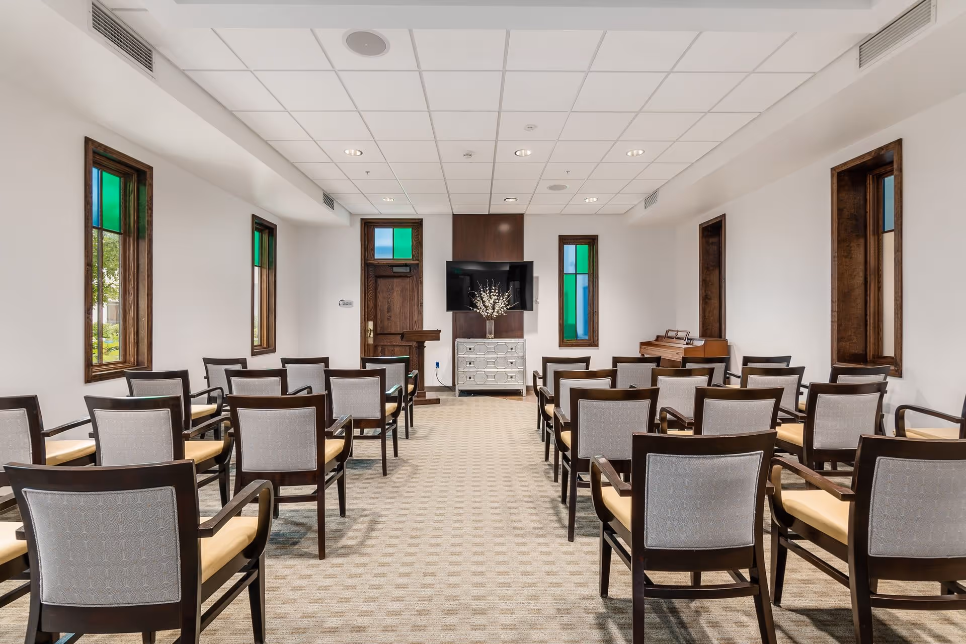 A small meeting or presentation room with rows of wooden chairs with beige cushions facing a flat-screen TV mounted on a wooden panel above a decorative chest. The room has white walls, a beige carpet, and several narrow windows with stained glass accents. A wooden podium and a piano are also visible at the front of the room.