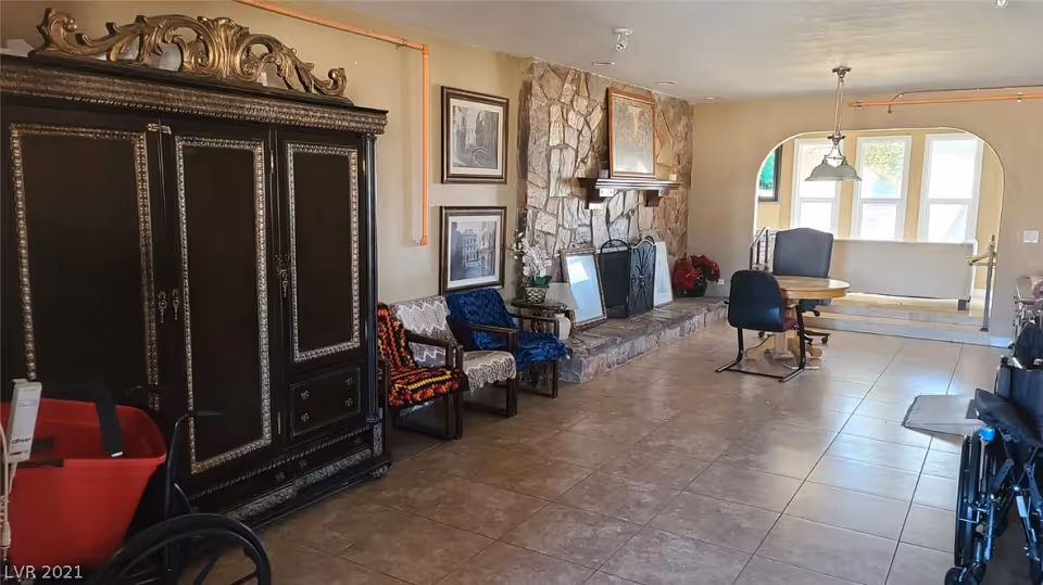 A spacious living area with a large ornate black cabinet on the left, three chairs with crocheted covers, a stone fireplace with framed pictures resting on the hearth, a round wooden table with two chairs, and large windows letting in natural light. The floor is tiled, and there is a wheelchair partially visible in the foreground.
