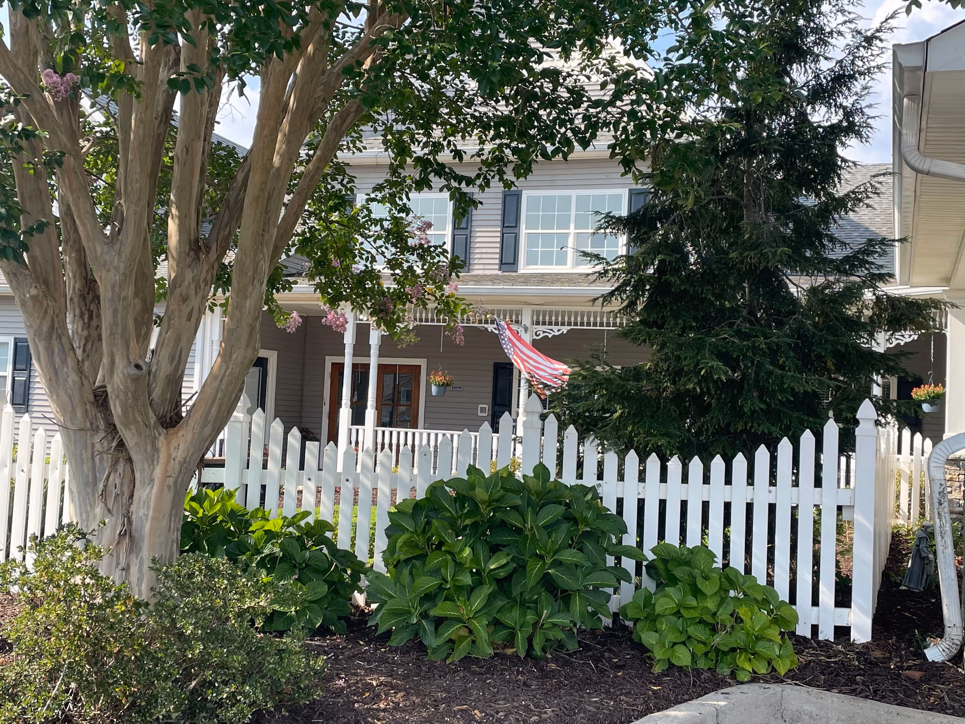 Front exterior of a house with a white picket fence, porch, American flag, and surrounding trees and shrubs.