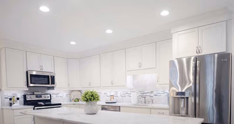 Bright modern kitchen with white cabinets, stainless-steel refrigerator and oven, and a marble island topped with a small potted plant.