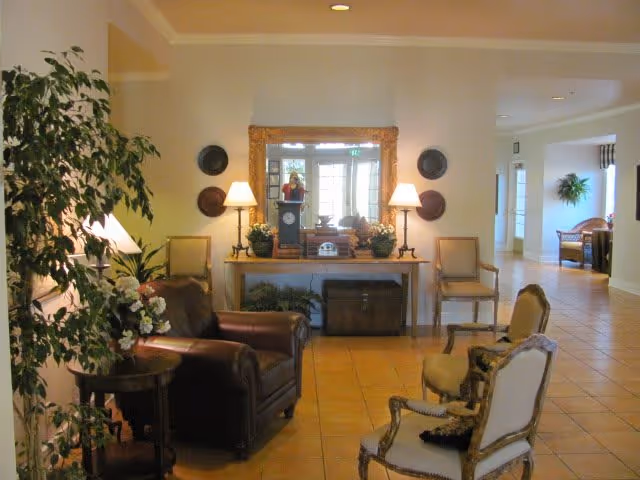 A cozy sitting area in a memory care community featuring a leather armchair, two upholstered chairs with cushions, a wooden side table with a floral arrangement, and a console table with two lamps, plants, decorative plates on the wall, and a large mirror reflecting the entrance door.
