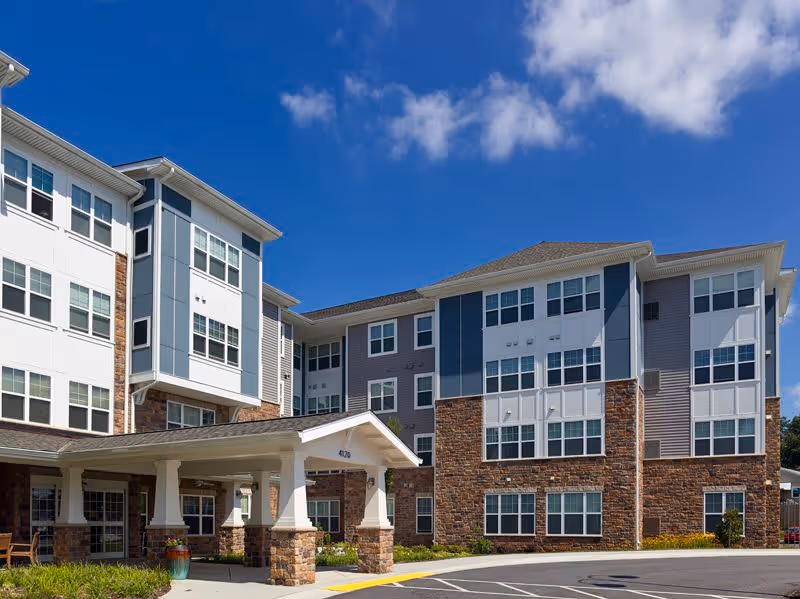 Exterior view of a multi-story senior living facility building with a covered entrance, large windows, and a mix of stone and siding on the facade under a blue sky with some clouds.