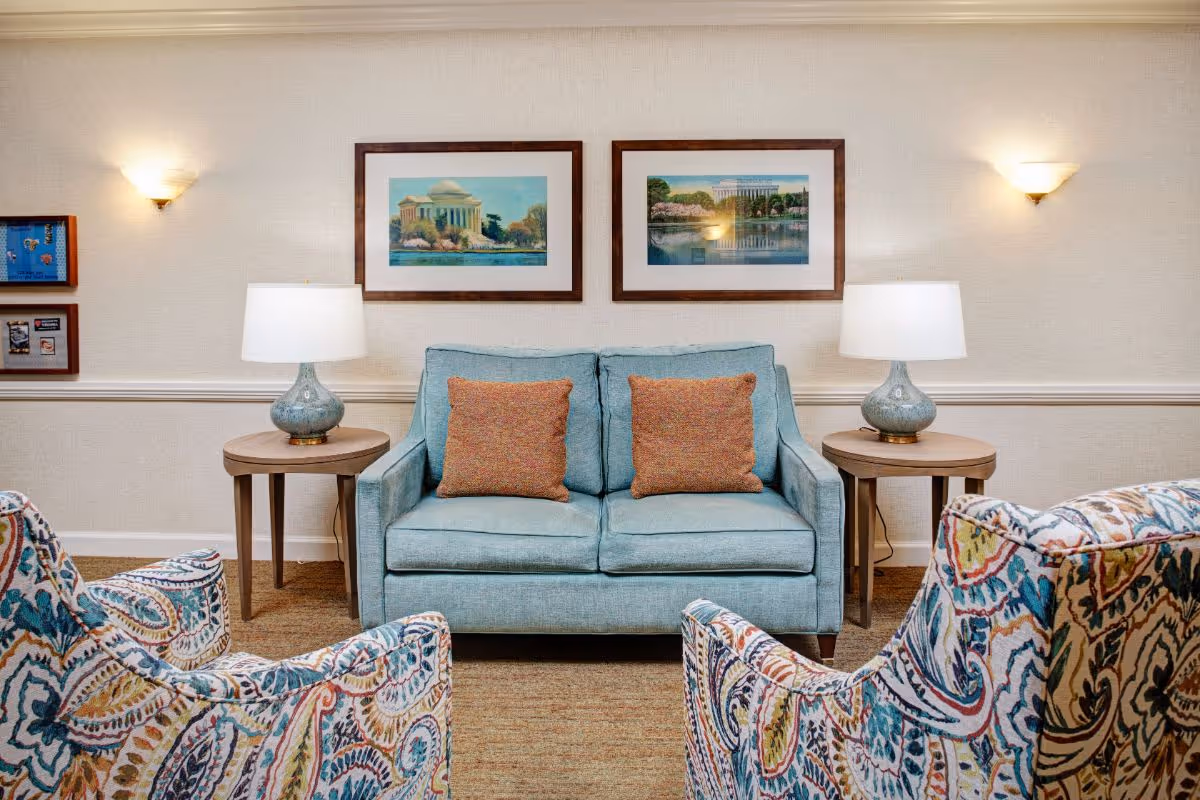 A cozy living room area with a light blue loveseat featuring two orange throw pillows, flanked by two wooden side tables each with a ceramic lamp. Two colorful patterned armchairs face the loveseat. The wall behind has two framed paintings of scenic buildings and two wall sconces providing warm lighting.
