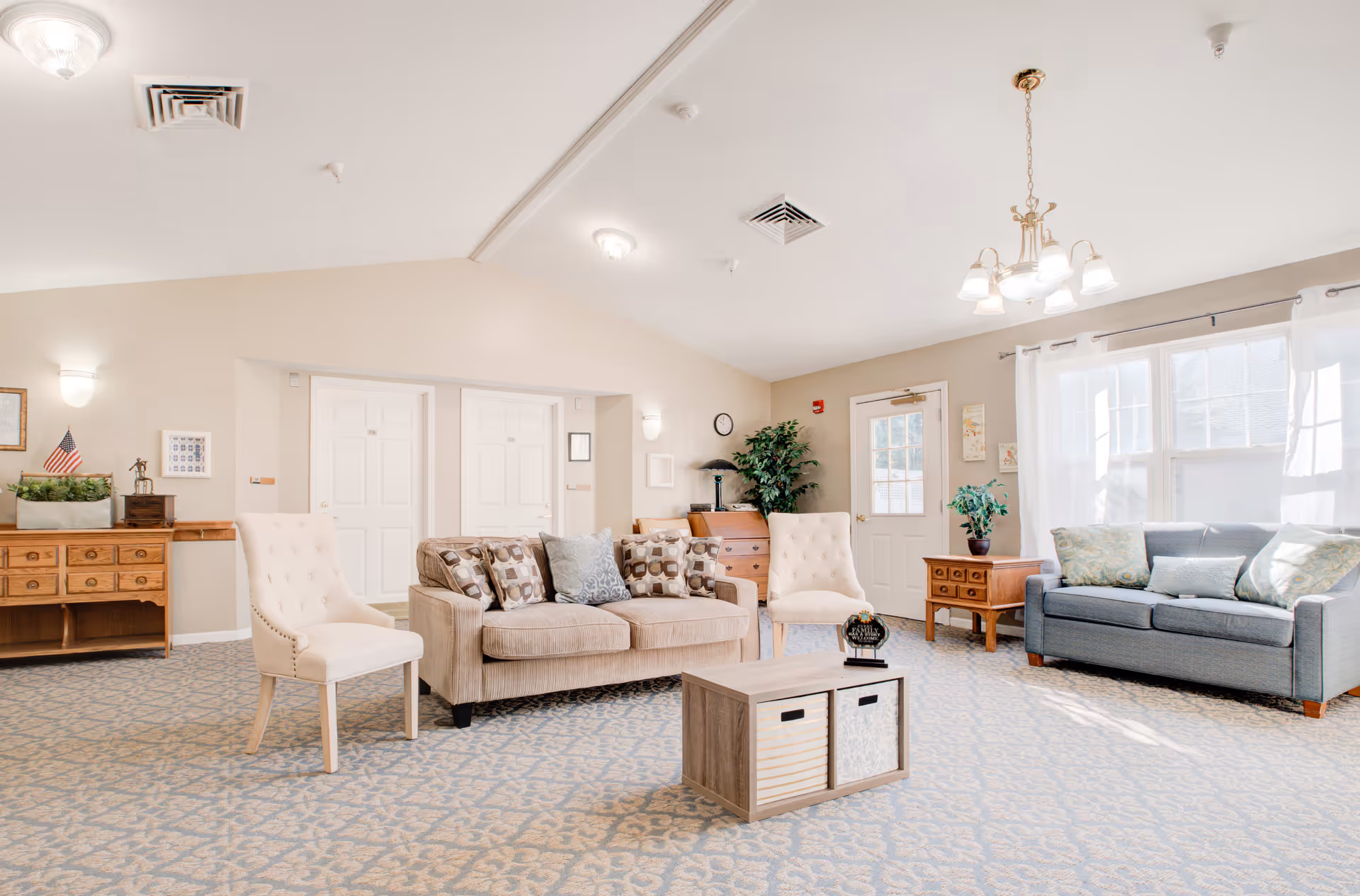 A bright and spacious living room area in a senior living facility with beige walls and patterned carpet. The room features a beige sofa with patterned cushions, two white upholstered chairs, a wooden coffee table with storage baskets, a blue sofa with cushions near a window with white curtains, and wooden side tables with plants and decorative items. The ceiling has recessed lighting and a chandelier, and there are double doors and a single door leading to other areas.