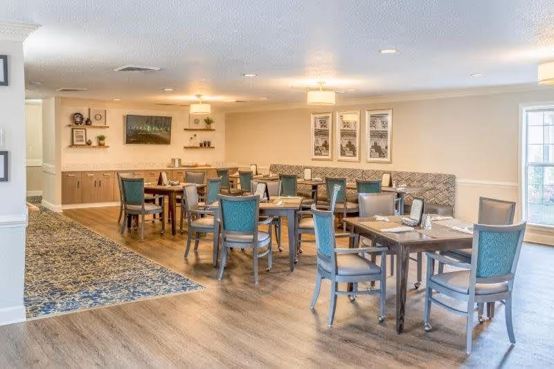 A bright and spacious dining room in a senior living facility with several wooden tables and chairs arranged neatly. The room features a patterned carpet area, light-colored walls, framed artwork, and a large window letting in natural light. There is a TV mounted on the wall and shelves with decorative items.