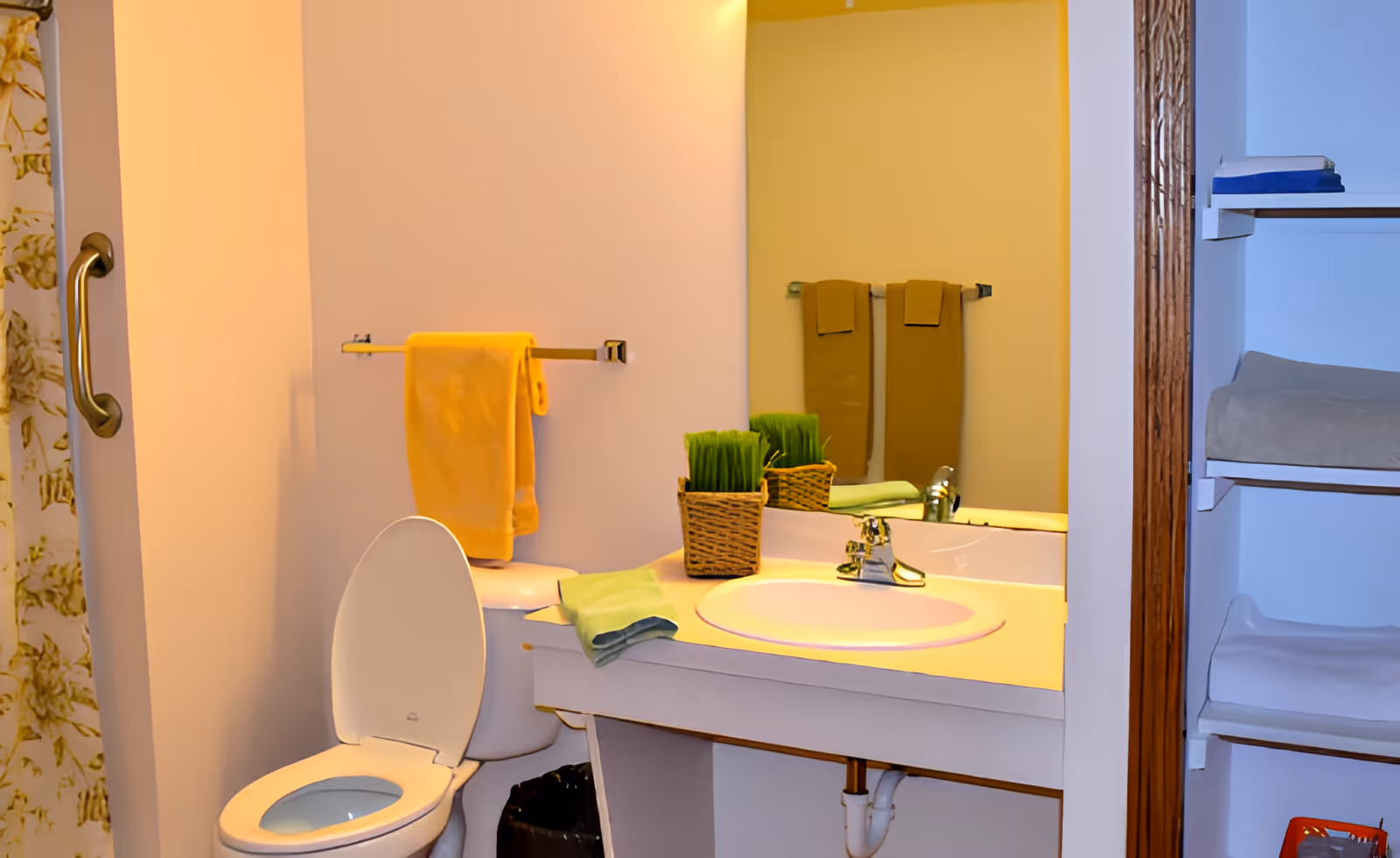 Small bathroom with an open toilet, sink vanity and mirror, yellow towels, and shelving to the right.