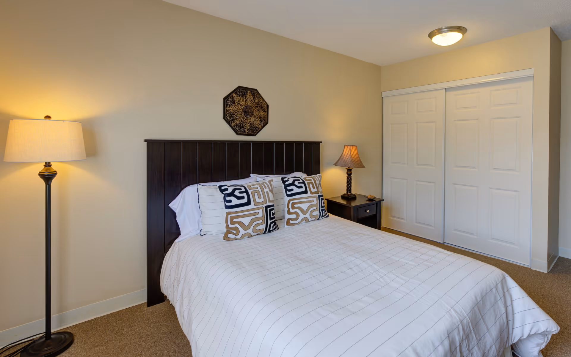 A neatly made bed with a dark wooden headboard, white bedding with subtle stripes, and four decorative pillows with geometric patterns. There is a floor lamp on the left side and a small bedside table with a lamp on the right side. Behind the bed is a beige wall with a decorative wall hanging, and to the right is a closet with white sliding doors.
