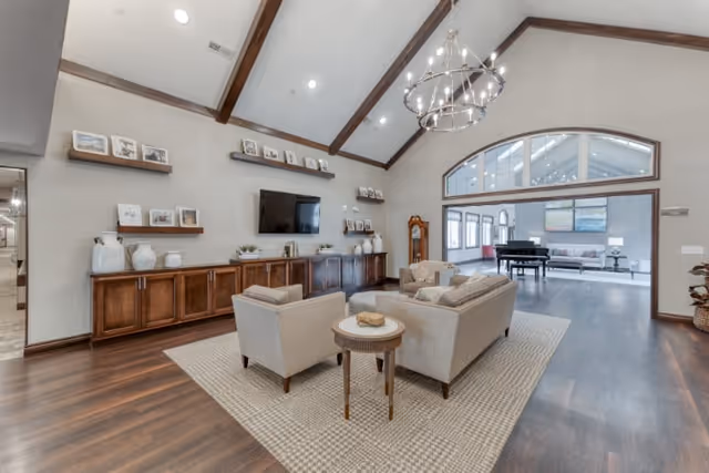 Vaulted common living room with sofas and chairs arranged on a rug, a wall-mounted TV and shelving, and a piano visible in the adjoining area.