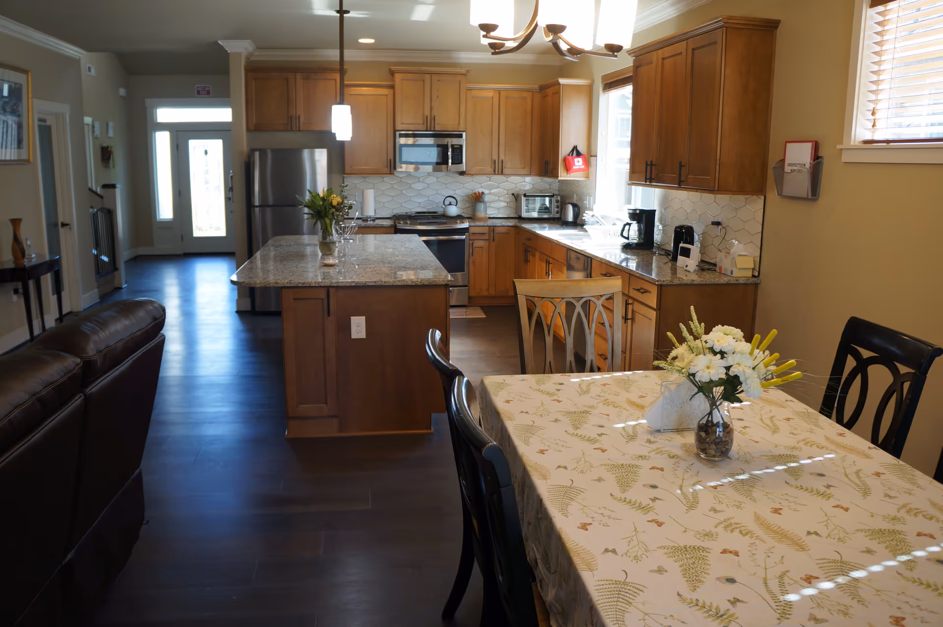 Interior view of a kitchen and dining area in a senior living facility. The kitchen features wooden cabinets, a stainless steel refrigerator, stove, microwave, and granite countertops. A kitchen island with a vase of flowers is in the center. The dining table is covered with a floral tablecloth and has a vase with white and yellow flowers. The room is well-lit with natural light coming through a window with blinds.