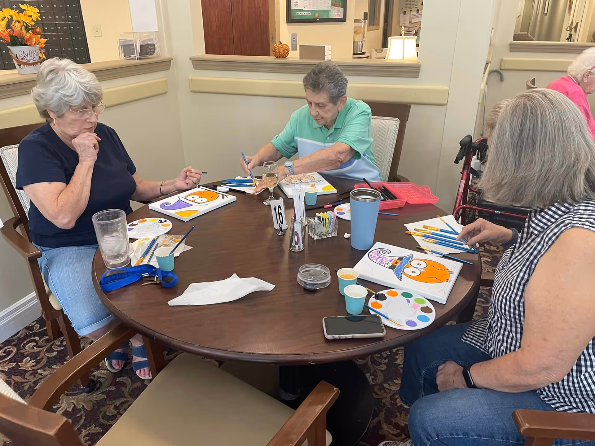 Three elderly women sitting around a round wooden table engaged in painting Halloween-themed pictures, including pumpkins and witches' hats. The table has paint palettes, brushes, cups, and a smartphone. The room has beige walls and a carpeted floor, with some decorations and a walker visible in the background.