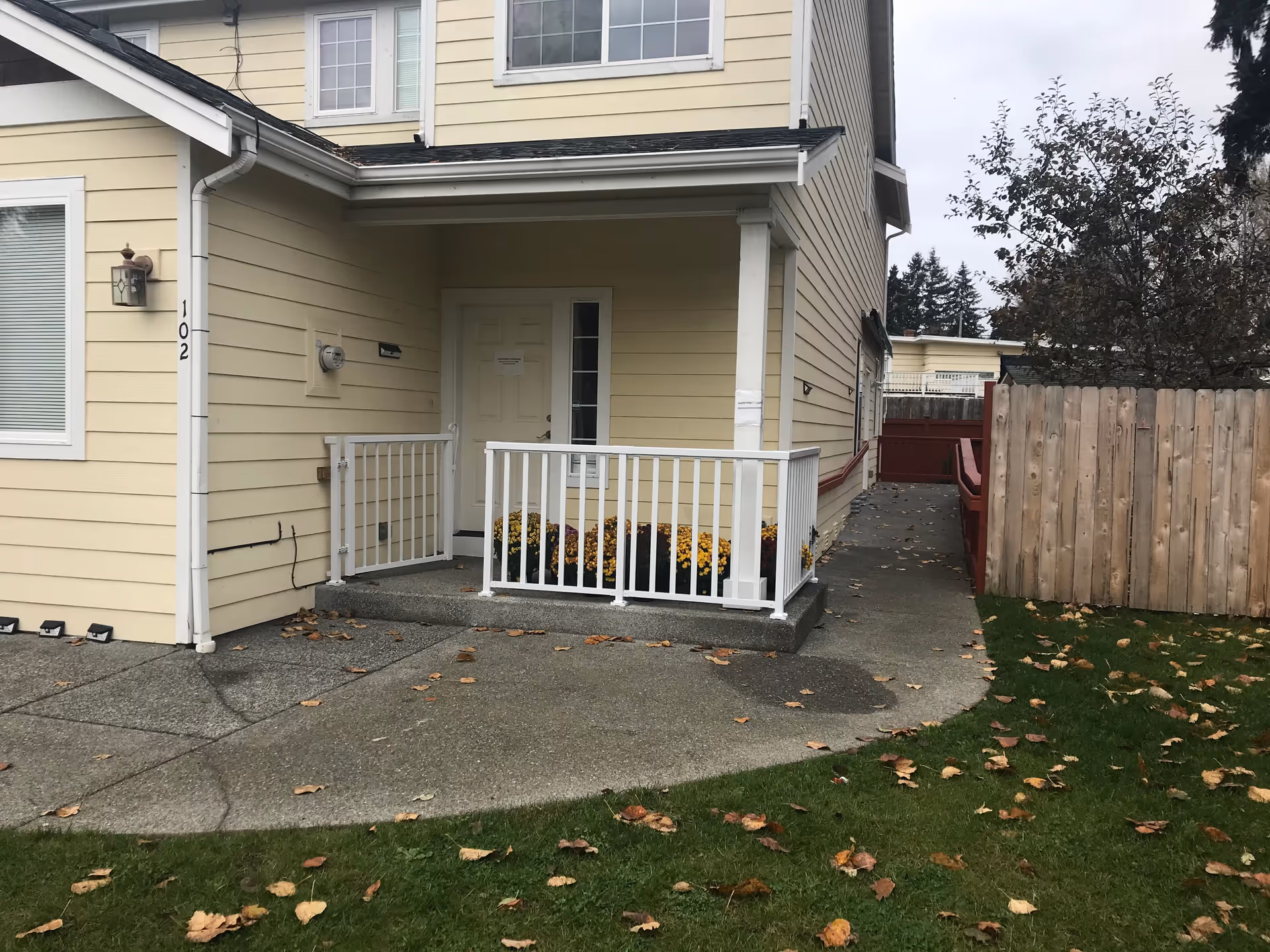 Exterior view of a yellow two-story building with white trim, featuring a small concrete porch with white railings and a door. There are yellow and purple flowers planted in front of the porch. A concrete pathway leads to a wooden fence and a red gate on the right side. The ground is covered with green grass and scattered fallen leaves.