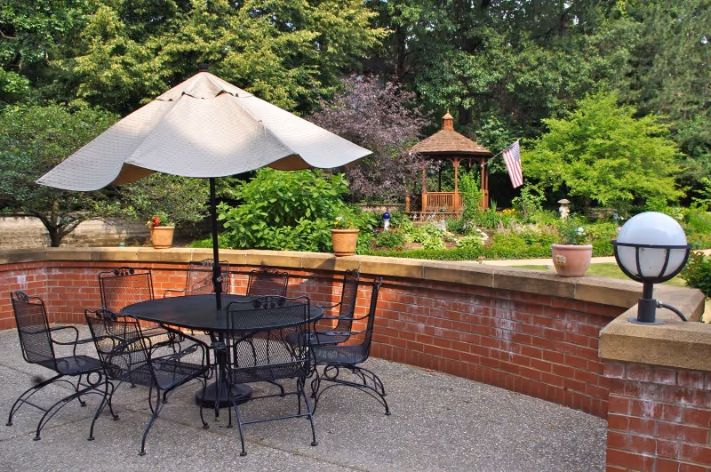 Outdoor patio area with a black metal table and six matching chairs under a large beige umbrella. The patio is enclosed by a low red brick wall with potted plants on top. In the background, there is a wooden gazebo surrounded by lush green trees and shrubs, with an American flag hanging nearby.