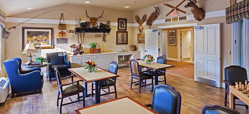 A cozy common area with several tables and chairs arranged on a wooden floor. The walls are decorated with rustic items including mounted deer heads, vintage snowshoes, fishing rods, and framed pictures. There is a small kitchenette area with cabinets and a sink, and the room is warmly lit with recessed ceiling lights. A chessboard is visible on a table in the foreground.