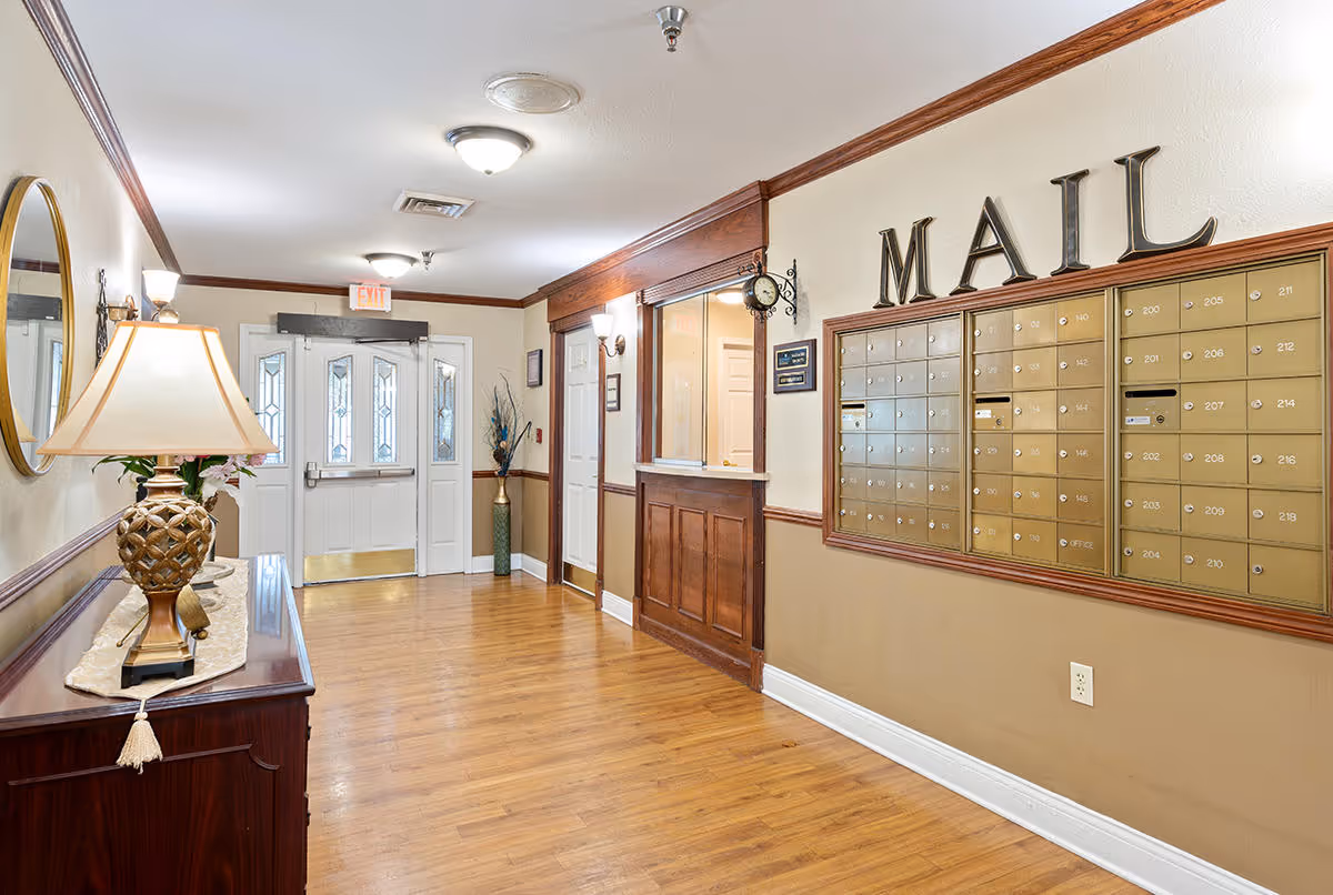 Interior hallway of a senior living facility with wooden flooring and beige walls. On the right side, there is a large mail area with multiple mailboxes labeled with numbers and the word 'MAIL' above them. On the left side, there is a wooden console table with a decorative lamp and a round mirror above it. At the end of the hallway, there is a white door with glass panels and an exit sign above it. The hallway is well-lit with ceiling lights and wall sconces.