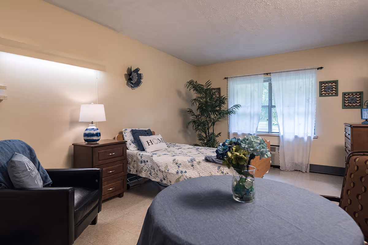 Furnished bedroom with a single bed, nightstand and lamp, armchair, small round table with flowers, and a window with sheer curtains.