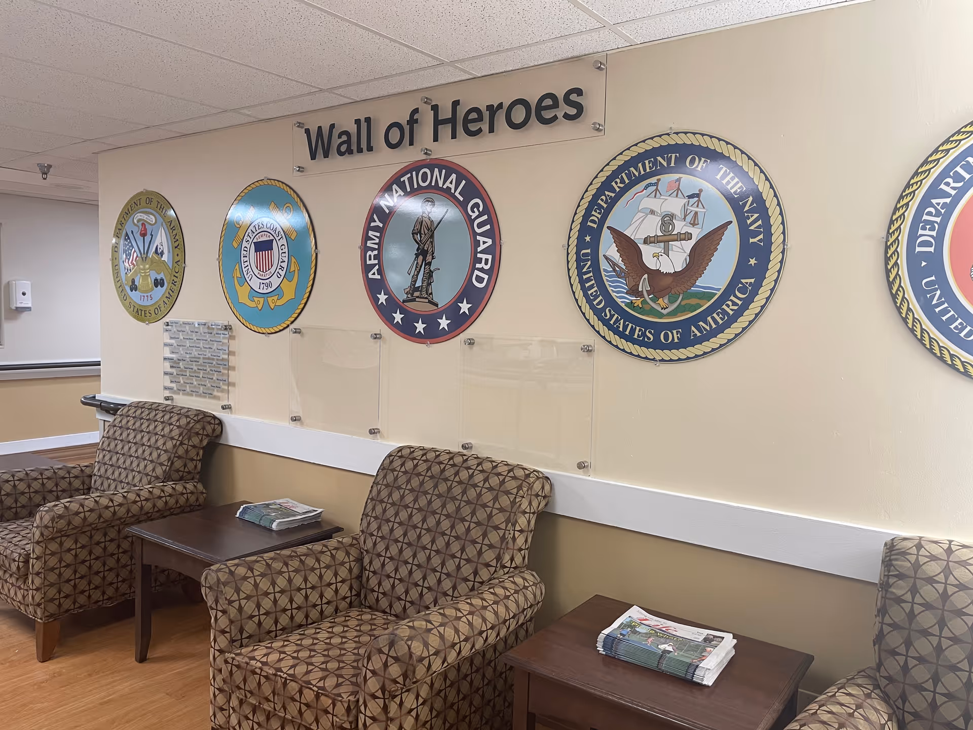 Seating area with patterned armchairs and side tables under a 'Wall of Heroes' display of military seals on the wall.