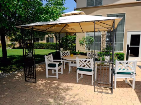 Shaded outdoor patio with a canopy covering a white table and chairs on a brick paver area beside a light-colored building.