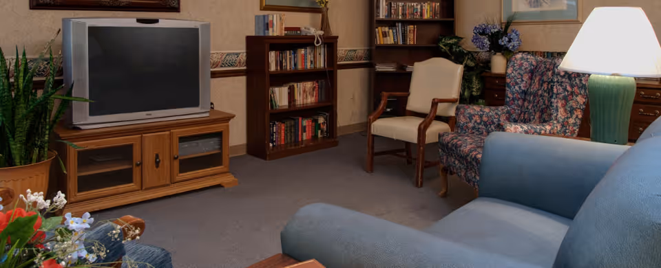 Cozy common area with a TV on a wooden stand, bookshelves, a floral armchair, and upholstered seating.