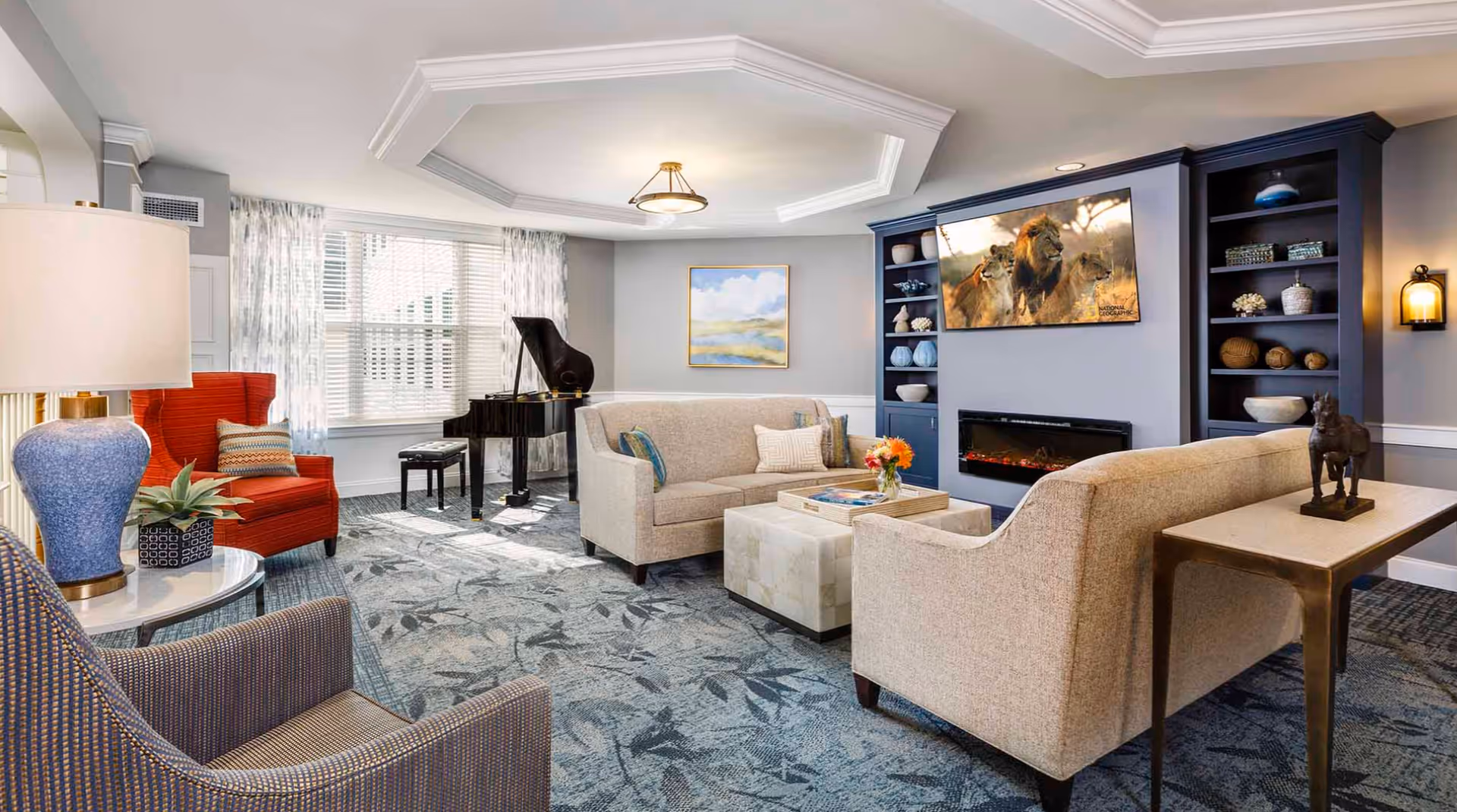 A bright and elegant living room with a patterned blue carpet, beige sofas, a red armchair, and a blue armchair. There is a grand piano near the window with white blinds and sheer curtains. A flat-screen TV mounted above a modern electric fireplace displays an image of lions. Built-in blue shelves flank the fireplace, holding decorative items. A small table with a horse sculpture is behind one of the sofas, and a ceiling light fixture is centered in a tray ceiling.