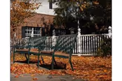 Two green metal benches on a sidewalk covered with fallen autumn leaves, with a white picket fence and a brick building partially visible in the background.