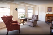 A cozy living room with two armchairs, one red patterned and one gray recliner, positioned near two large windows with white blinds. A small wooden table with a flower vase sits between the chairs. In the background, there is a wooden desk with a chair and a framed picture on the wall.