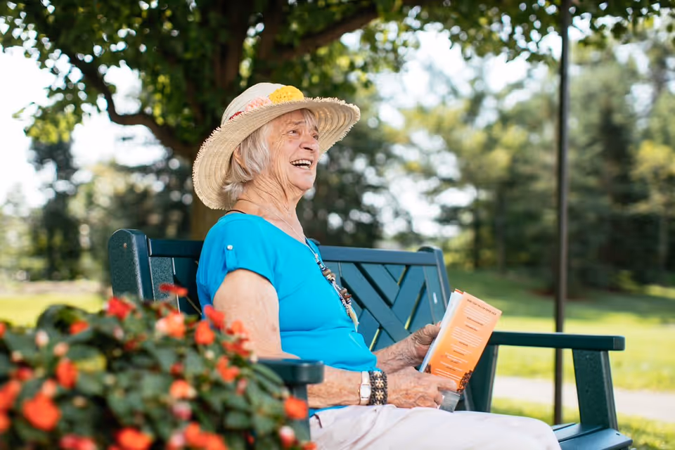 An elderly woman wearing a straw hat with a flower decoration and a blue shirt is sitting on a green bench outdoors, holding a book and smiling. There are red flowers in the foreground and trees in the background under a sunny sky.