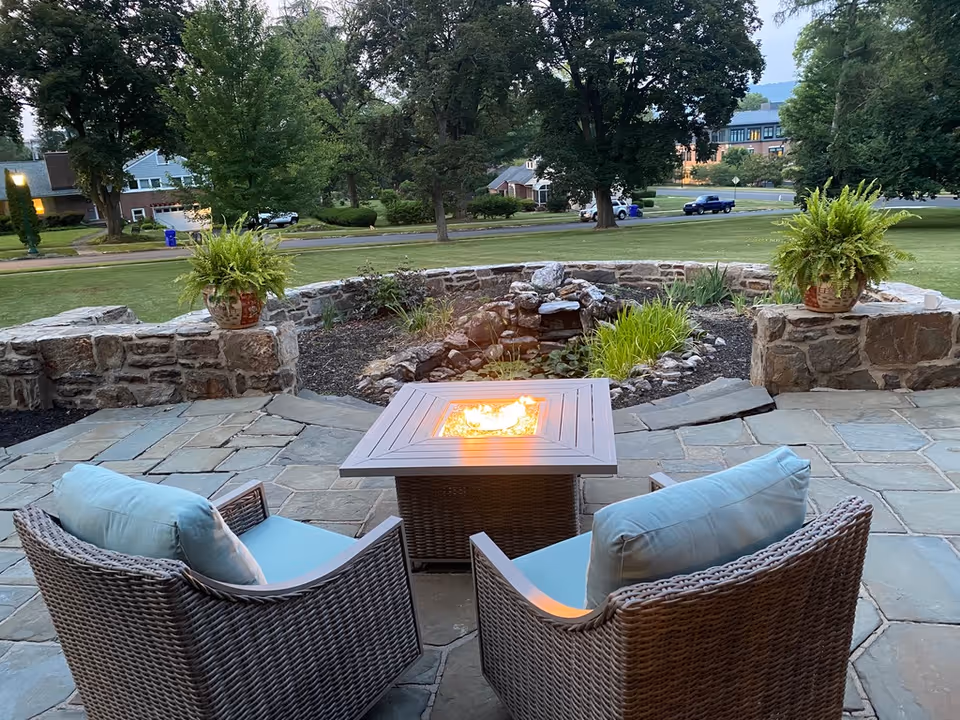 Stone patio with two wicker chairs facing a lit square fire pit table overlooking a small pond and grassy lawn.