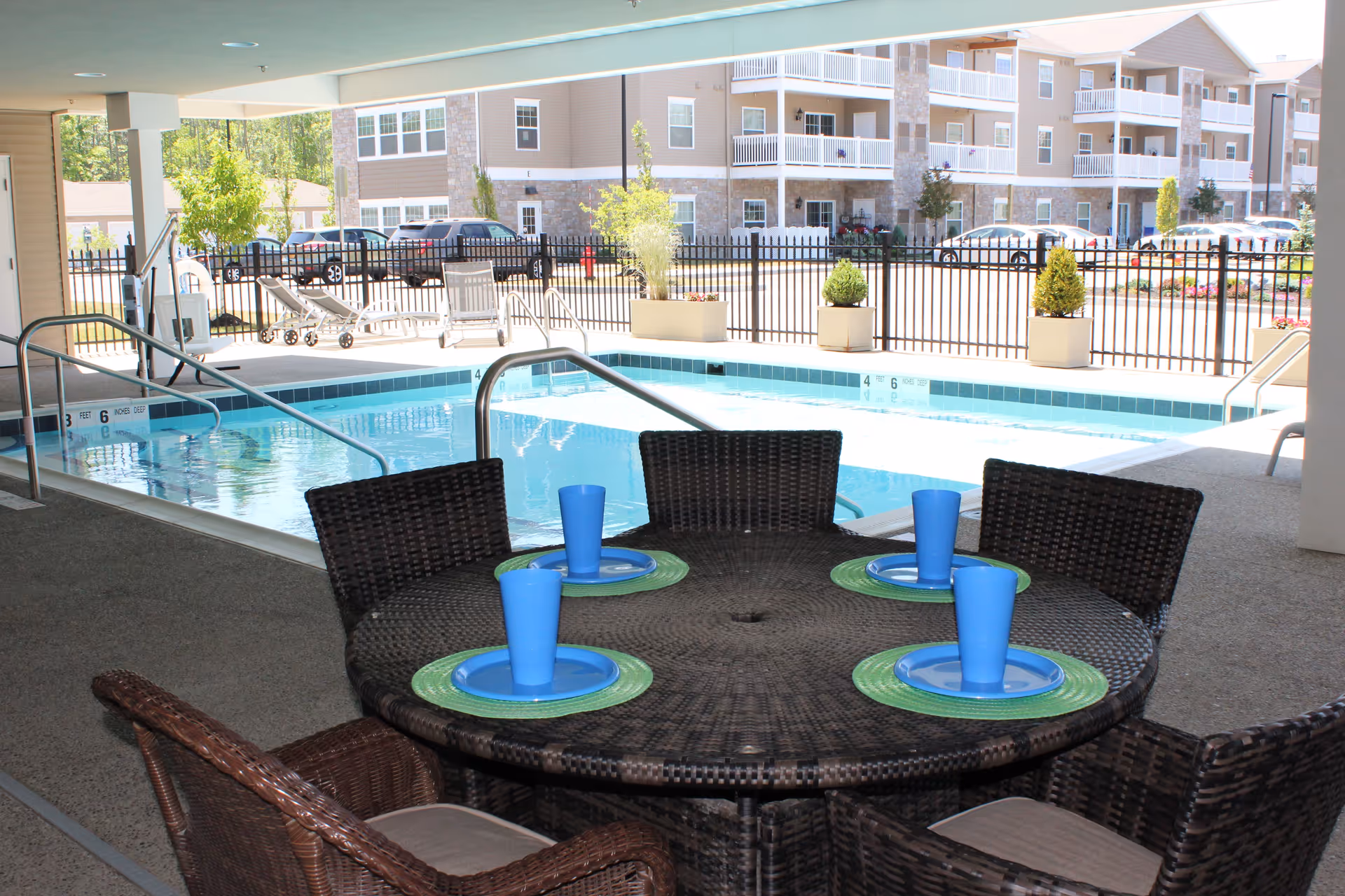 Wicker patio table with blue cups and plates beside a covered outdoor pool with apartments visible beyond.